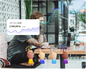 Homem com tatuagens a trabalhar num computador portátil num café ao ar livre, com um gráfico de vendas por SMS e ícones emoji de mãos levantadas sobrepostos, ilustrando um maior envolvimento.